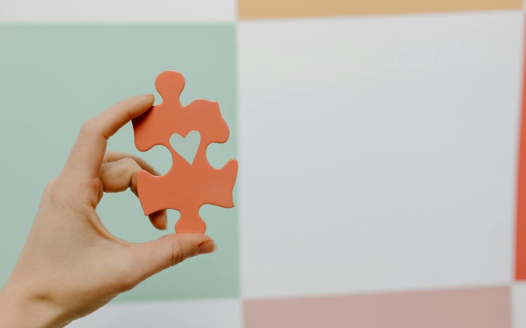 Close-up of a hand holding a heart-shaped puzzle piece against a colorful background.